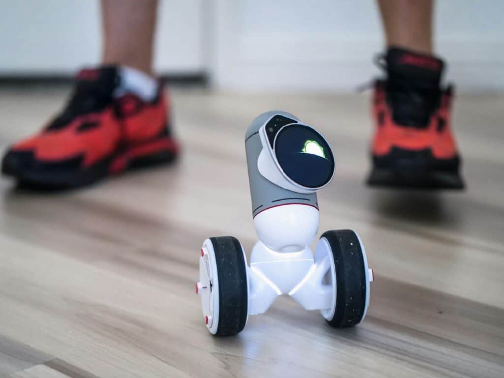 Close-up of a two-wheeled mini robot on a wooden floor with sneakers in background.