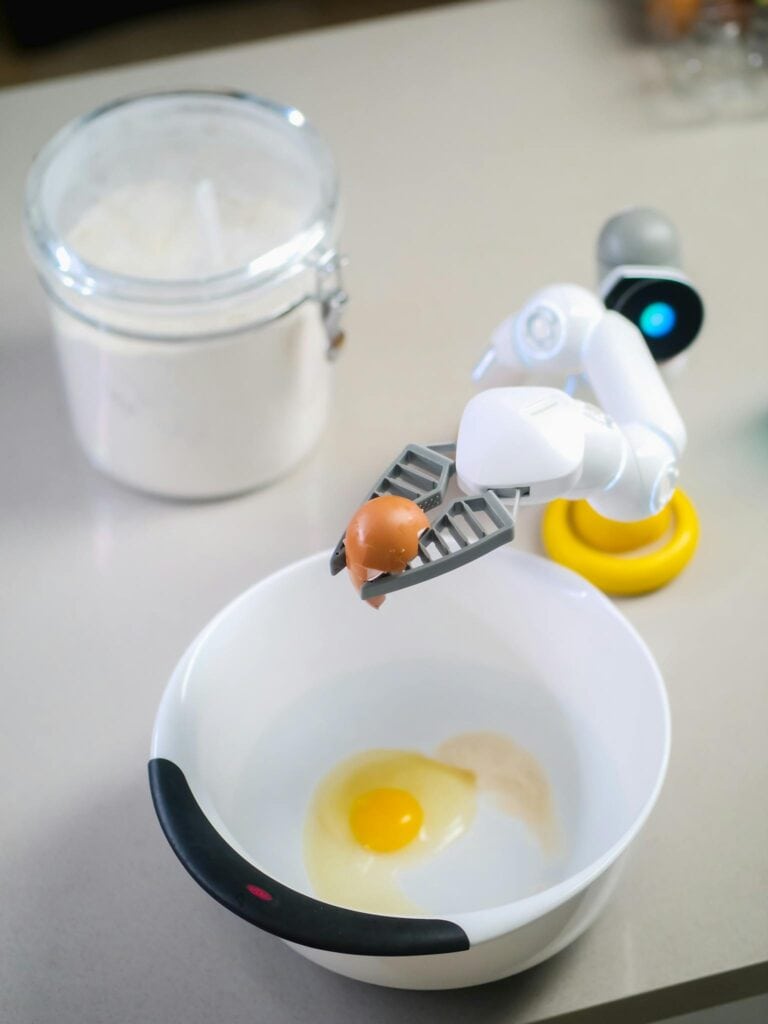 Futuristic robotic arm with egg over bowl prepares ingredients in modern kitchen.