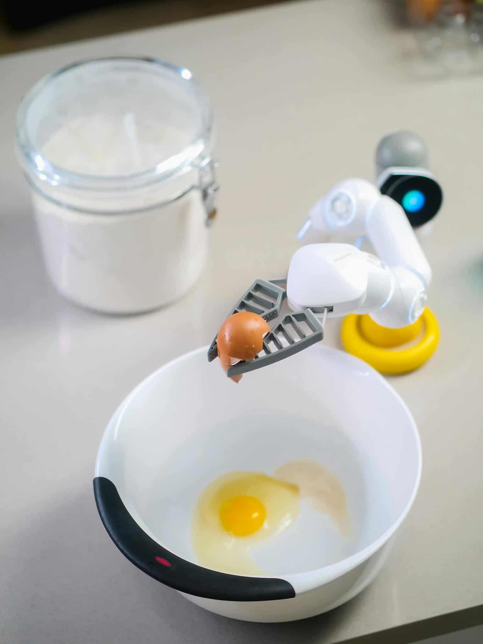 Futuristic robotic arm with egg over bowl prepares ingredients in modern kitchen.